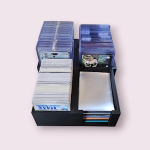 May include: Black card storage organizer with multiple compartments for holding trading cards. The organizer has a clear plastic top for viewing the cards and a silver compartment for storing card sleeves.