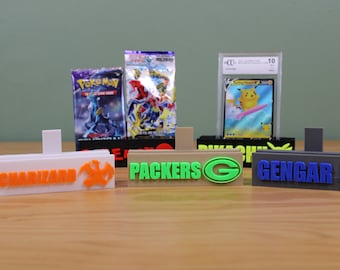 May include: Five 3D printed card holders with different designs. The holders are white, beige, green, gray, and blue. The holders have the words "CHARIZARD", "PACKERS", "GENGAR", and "PIKACHU" printed on them.