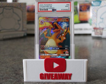 May include: A white plastic stand with a red play button and the word "GIVEAWAY" printed on it. A graded Pokemon card featuring Charizard GX is displayed on the stand. The card is in a protective case with a PSA label that reads "#60 MINT 9 61040623".