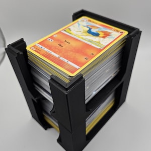 May include: Black plastic card holder with a stack of Pokemon trading cards. The top card features a red and yellow image of a bird-like Pokemon called "Charizard" with the text "Tackle" and "Flare" on the card.