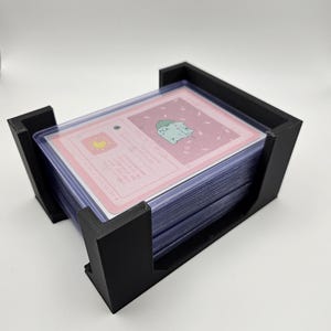 May include: A black plastic card holder with a stack of clear plastic card sleeves. The cards have a pink background with a light blue illustration of a cat.