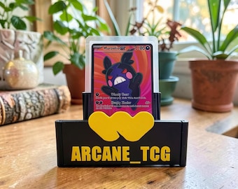 May include: A black plastic trading card display stand featuring a yellow heart-shaped accent and the text ARCANE_TCG in yellow lettering. The stand holds a Morpeko GX trading card encased in a clear protective sleeve with a pink and black design.