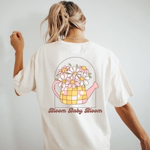 Puede incluir: Camiseta blanca con un gráfico de una regadera rosa llena de margaritas blancas. El texto "Bloom Baby Bloom" está impreso debajo del gráfico.