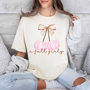 May include: A cream colored t-shirt with a pink and brown pumpkin design and the text "a fall girly".
