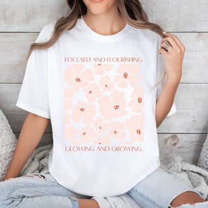Pode incluir: Camiseta branca com um design floral rosa e o texto "Focused and Flourishing. Glowing and Growing."