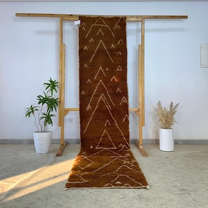 Puede incluir: Una alfombra larga y rectangular de color marrón con un diseño geométrico de triángulos y líneas blancas. La alfombra se exhibe en un estante de madera contra una pared blanca. La alfombra está hecha de un material texturizado.