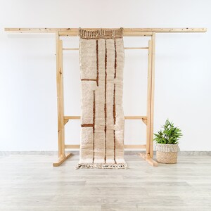 May include: A beige and brown striped rug is displayed on a wooden rack. The rug has vertical brown stripes and a fringed edge. The rack is made of light-colored wood and is set against a white wall. A small potted plant sits to the right.