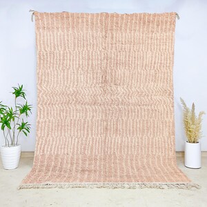 May include: A large, rectangular rug in a soft, muted pink hue. The rug features a textured, vertical striped pattern across its surface. The rug is displayed against a white wall, with a fringe along the bottom edge. The rug is a decorative home decor item.