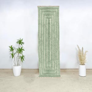 May include: A rectangular, light green rug with a geometric design of vertical and horizontal lines. The rug has a soft, textured appearance and is displayed against a white wall. The rug is approximately 60 inches long.