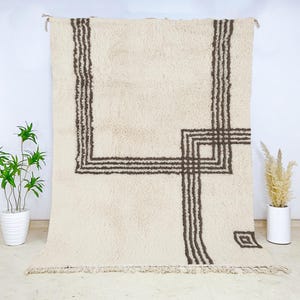 May include: Cream-colored, rectangular rug with a geometric design in dark brown. The rug features a central square and vertical lines, with a fringe along the bottom edge. The rug is displayed against a white wall, showcasing a minimalist aesthetic.