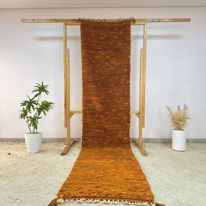 May include: A long, rectangular, orange and brown rug with a textured surface and fringe detail. The rug is displayed on a wooden rack, showcasing its length and design. The rug is approximately 10 feet long.