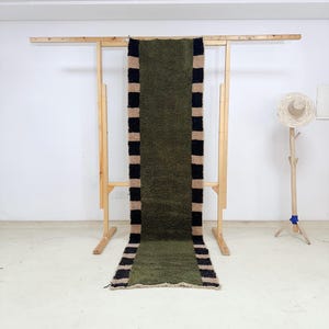 Puede incluir: Una alfombra larga de color verde oliva con bordes a rayas negras y beige. La alfombra se exhibe en un marco de madera, resaltando su forma rectangular y superficie texturizada. El diseño de la alfombra añade un toque moderno.