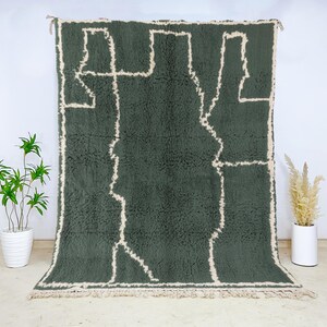 May include: A large, rectangular, dark green rug with a cream-colored geometric design. The rug has a shaggy texture and a fringe along the bottom edge. The rug is displayed against a white wall.