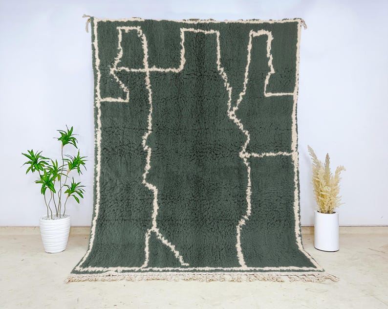 May include: A large, rectangular, dark green rug with a cream-colored geometric design. The rug has a shaggy texture and a fringe along the bottom edge. The rug is displayed against a white wall.