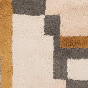 May include: A close-up of a textured rug with a geometric pattern. The rug features blocks of cream, gray, pink, and mustard yellow. The rug's design is modern and abstract, suitable for home decor.