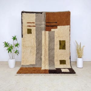 May include: A rectangular area rug featuring a high-pile texture with a geometric pattern in shades of cream, brown, gray, and olive green. The design includes vertical bands and square blocks, finished with a short fringe along the bottom edge.