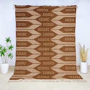 Op de afbeelding: Een bruin en beige patroonkleed met een herhalend pijl- en bladerontwerp. Het kleed heeft een gefranjerde rand en wordt tegen een witte muur getoond. Het kleed is een decoratief huishoudtextiel.