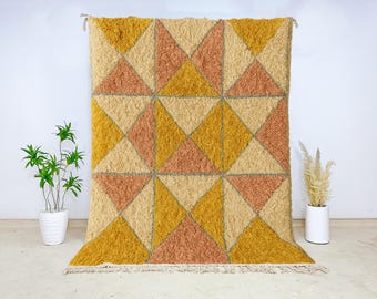 May include: A large, textured rug with a geometric pattern of triangles in shades of gold, peach, and beige. The rug has a fringe along the bottom edge and is displayed against a white wall. The rug is a decorative home accent.