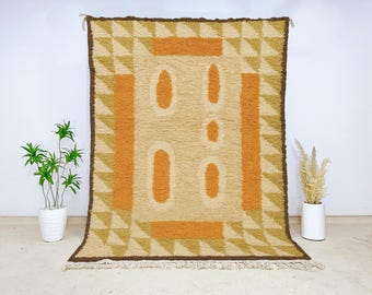 May include: A rectangular, beige rug with an abstract design. The rug features a central panel with four vertical ovals and two horizontal rectangles in orange. The border is brown, and the corners have triangles in shades of beige and orange. The rug has a fringe.