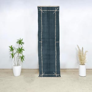 May include: A rectangular, teal-colored rug with a cream-colored geometric border design. The rug has a fringed edge and is displayed against a white wall. The rug is a decorative home accent.