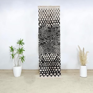 Puede incluir: Una alfombra de pasillo con estampado en blanco y negro con un diseño geométrico. La alfombra presenta un patrón de puntos repetidos y un diseño central en forma de estrella. La alfombra es rectangular y tiene un borde con flecos. La alfombra se exhibe contra una pared blanca.