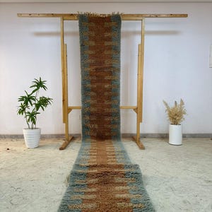 Könnte beinhalten: Ein langer, rechteckiger Teppich mit einem geometrischen Muster in Braun- und Blaugrüntönen. Der Teppich wird auf einem Holzgestell präsentiert, das seine Länge und Textur zeigt. Quasten verzieren die Enden des Teppichs und fügen ein dekoratives Element hinzu.