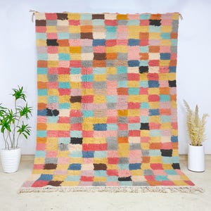 May include: A colorful, rectangular rug with a checkerboard pattern. The rug features squares of various colors, including pink, blue, yellow, and brown. The rug has a fringe along the bottom edge and is hanging against a white wall.