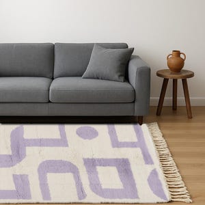 May include: A cream-colored rug with a geometric pattern in lavender. The rug is rectangular with a fringed edge and is placed in front of a gray sofa. A small wooden side table and a decorative vase are also visible.