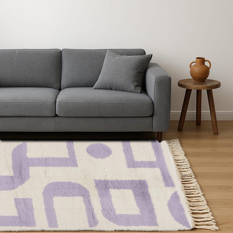 May include: A cream-colored rug with a geometric pattern in lavender. The rug is rectangular with a fringed edge and is placed in front of a gray sofa. A small wooden side table and a decorative vase are also visible.