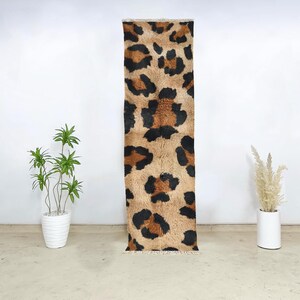May include: A rectangular rug with a leopard print design. The rug features a tan background with black and brown spots. The rug is displayed vertically against a white wall, with a white pot of plants on either side.