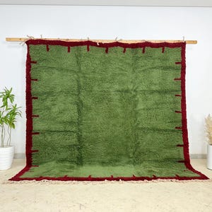 May include: A large, rectangular green rug with a deep red border and fringe. The rug is displayed on a wooden rod against a white wall. The rug's texture appears soft and plush, suitable for a living room or bedroom.