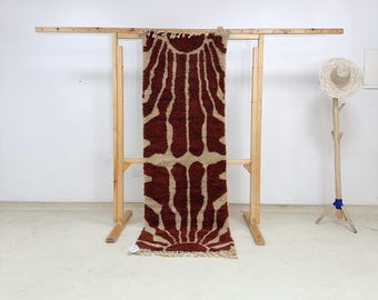 May include: A rectangular rug with a sunburst pattern in brown and beige tones. The rug is displayed on a wooden rack, highlighting its texture and design. The rug has a fringed edge. The rug is approximately 150 cm in length.