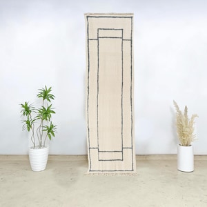 May include: A rectangular, cream-colored rug with a geometric design in dark gray. The rug has a fringe at the bottom and is displayed against a white wall. The rug's dimensions are not visible.