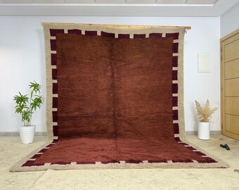 May include: A large, rectangular rug with a deep red center and a beige border. The border is accented with a repeating pattern of dark red rectangles. The rug is displayed against a white wall, showcasing its size and texture. The rug is a floor covering.