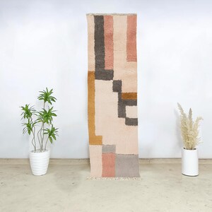 This hand-knotted Moroccan Beni Ourain runner rug showcases a harmonious geometric pattern in blush, mustard, grey, and cream wool. Woven by Berber women using traditional techniques, it features soft, color-blocked sections that bring warmth and structure to boho or modern interiors. The artisan-crafted texture and soothing palette make this wool runner ideal for entryways, hallways, or narrow spaces looking for both coziness and design impact.