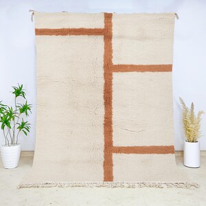 May include: Cream-colored, rectangular rug with a geometric design in a warm brown hue. The rug features a thick, textured pile and a fringe along the bottom edge. The design consists of intersecting lines creating a bold, modern look. The rug is displayed against a white background.