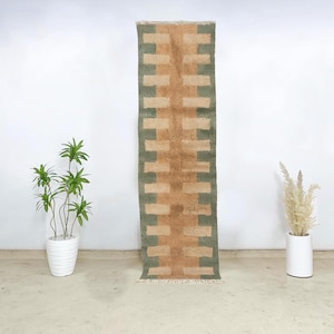 Peut inclure: Un tapis rectangulaire avec un motif géométrique dans les tons de beige, vert et marron. Le tapis a un motif en forme de briques et est affiché verticalement. Le tapis a un bord effiloché en bas.