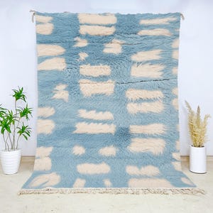 May include: A large, rectangular rug with a light blue background and cream-colored rectangular patterns. The rug has a shaggy texture and a fringe along the bottom edge. The rug is displayed against a white wall.