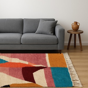May include: A colorful geometric rug with a fringed edge. The rug features a mix of pink, orange, blue, and cream shapes. A gray sofa and pillow are in the background, along with a small wooden side table and a brown vase.
