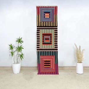 May include: A long, rectangular rug with a geometric pattern. The rug features three stacked squares in shades of red, black, green, and yellow. The rug has a fringed edge and is displayed against a white wall.