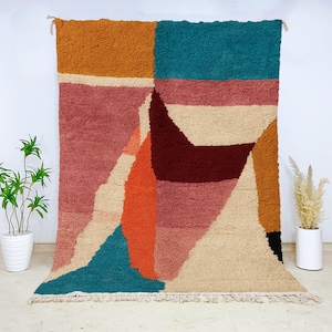 Artistic Moroccan wool rug with bold abstract shapes in teal, rust, blush pink, cream, and burgundy. Handwoven by skilled artisans using natural wool, this plush carpet blends contemporary art with Moroccan heritage. Ideal for boho, eclectic, or modern living rooms and bedrooms, it delivers vibrant energy, cozy texture, and a striking centerpiece for creative home interiors, combining comfort, craftsmanship, and artistic flair.