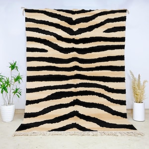 Op de afbeelding: Een groot, rechthoekig tapijt met een zebra streep patroon. Het tapijt heeft afwisselende zwarte en beige strepen, waardoor een gedurfd en grafisch ontwerp ontstaat. Het tapijt heeft een franje langs de onderrand en wordt tegen een witte muur getoond.