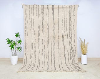 May include: Cream-colored rug with vertical dark gray stripes. The rug has a textured surface and a fringed edge. It is hanging against a white wall, with plants in white pots on either side.