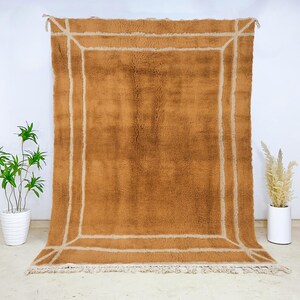 May include: A large, rectangular, brown rug with a cream-colored border. The rug has a thick, textured pile and a fringe along the bottom edge. The border is a simple, geometric design. The rug is displayed against a white wall.