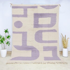 Minimalist Moroccan wool rug featuring a soft cream base with abstract geometric lavender shapes. This handwoven plush carpet combines modern simplicity with traditional Berber craftsmanship, crafted from premium natural wool. Ideal for Scandinavian, minimalist, or contemporary interiors, this soft rug adds a clean and calming touch to living rooms, bedrooms, or home offices, offering cozy texture, artistic elegance, and timeless Moroccan authenticity.