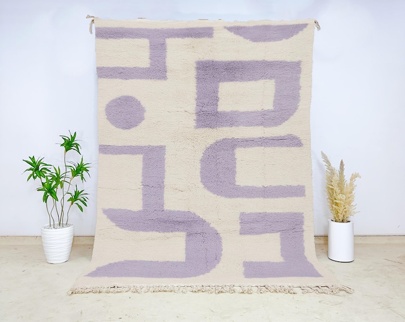 Minimalist Moroccan wool rug featuring a soft cream base with abstract geometric lavender shapes. This handwoven plush carpet combines modern simplicity with traditional Berber craftsmanship, crafted from premium natural wool. Ideal for Scandinavian, minimalist, or contemporary interiors, this soft rug adds a clean and calming touch to living rooms, bedrooms, or home offices, offering cozy texture, artistic elegance, and timeless Moroccan authenticity.
