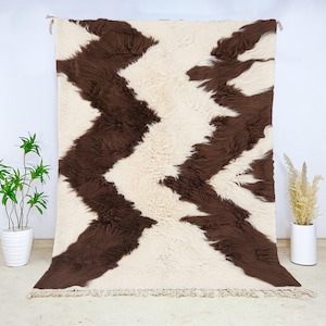 Puede incluir: Una alfombra rectangular grande con un fondo color crema y un llamativo patrón en zigzag en marrón oscuro. La alfombra tiene una textura peluda y flecos en el borde inferior. La alfombra se exhibe contra una pared blanca.