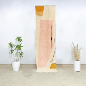 Puede incluir: Una alfombra rectangular de color crema con un diseño geométrico. La alfombra presenta un gran rectángulo rosa claro, cuadrados amarillos y líneas negras y naranjas. La alfombra se muestra verticalmente y tiene un borde con flecos.