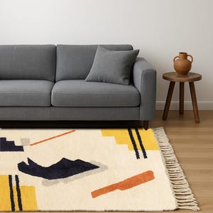 Puede incluir: Una alfombra de &aacute;rea color crema con un dise&ntilde;o geom&eacute;trico que presenta formas amarillas, azul marino y naranja. La alfombra tiene un borde con flecos y est&aacute; colocada frente a un sof&aacute; gris. Tambi&eacute;n se ven una peque&ntilde;a mesa auxiliar de madera y un jarr&oacute;n decorativo.