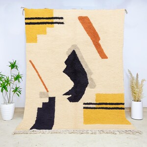 Hand-knotted Beni Ourain wool rug featuring a modern abstract design with mustard yellow, burnt orange, and black geometric shapes on a cream base. The plush texture and artisan craftsmanship make this Moroccan rug a perfect centerpiece for bohemian, modern, or eclectic interiors, suitable for living rooms, bedrooms, or studios seeking bold color and design.